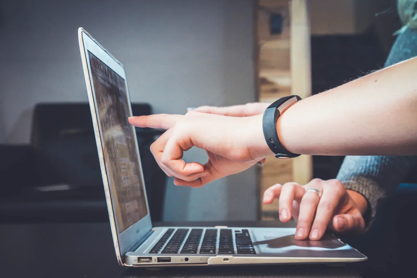 Side view of an open laptop on a dark table as one person points at the screen, wearing a black fitness band, while another person's hand wearing a ring rests on the trackpad.