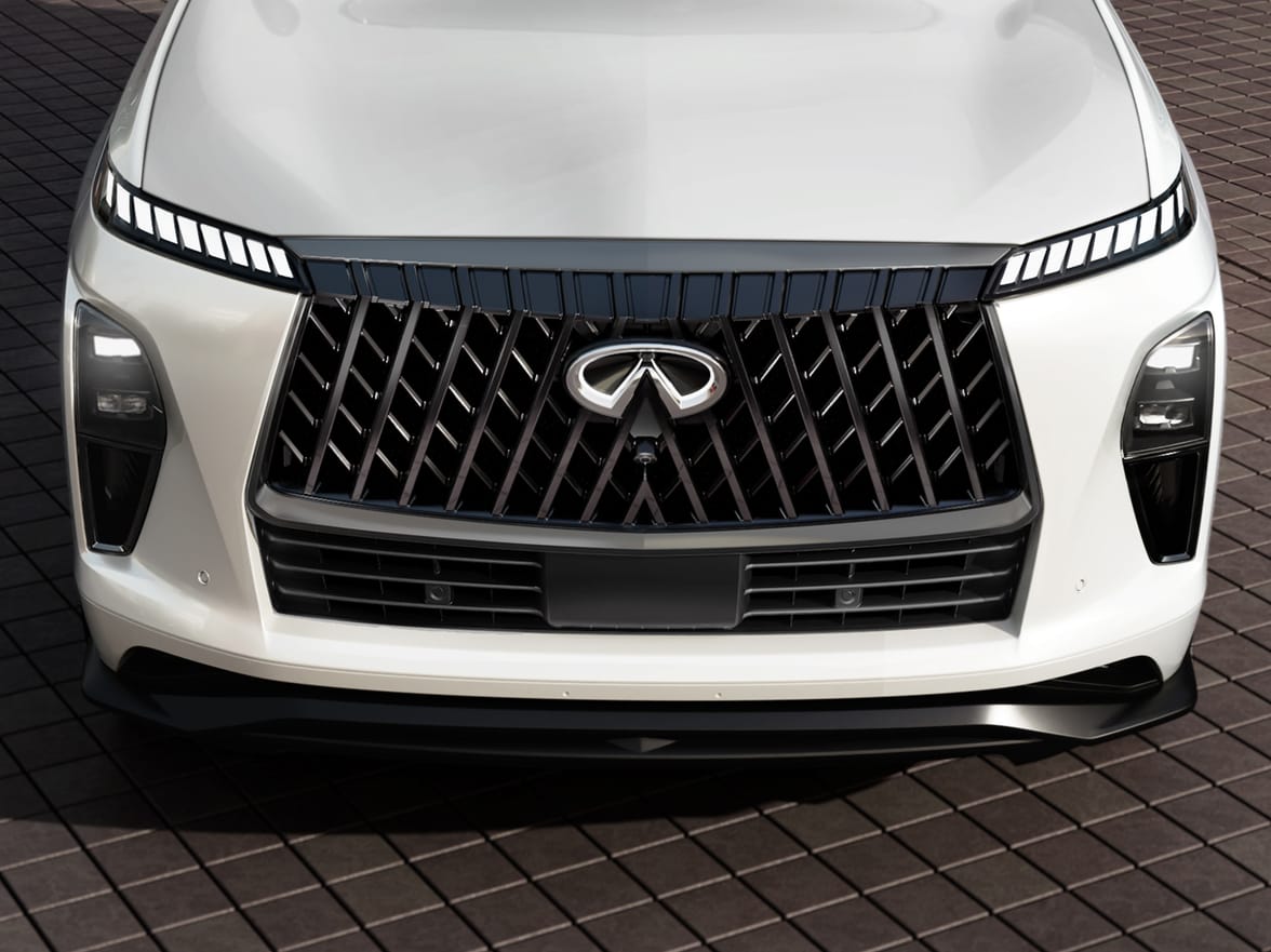 High-angle front close-up of a pearl white 2027 INFINITI QX80 SPORT showing the gloss-black double-arch grille, chrome emblem, and digital piano-key LED lights.