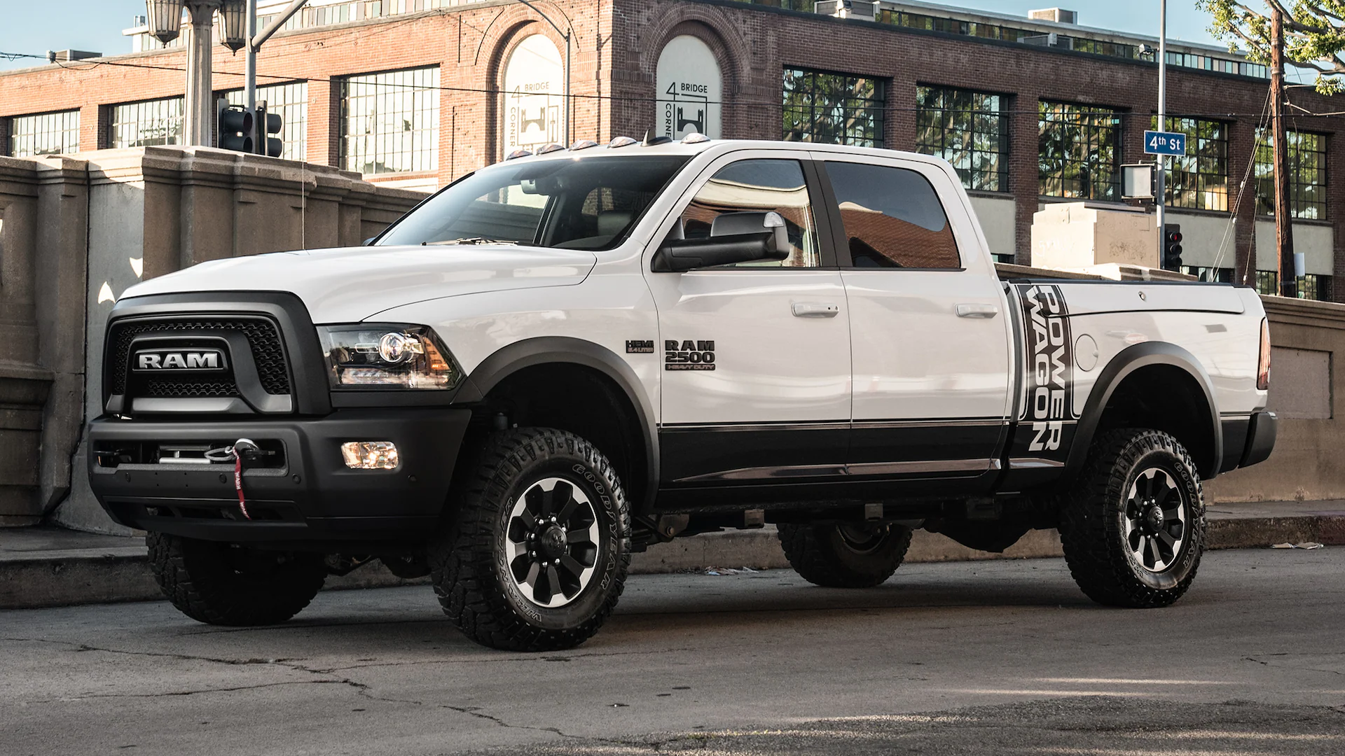 a white Ram 2500 in a front side view that has power wagon on the side of the front of the bed. The truck is parked on the side of the street in a city landscape.