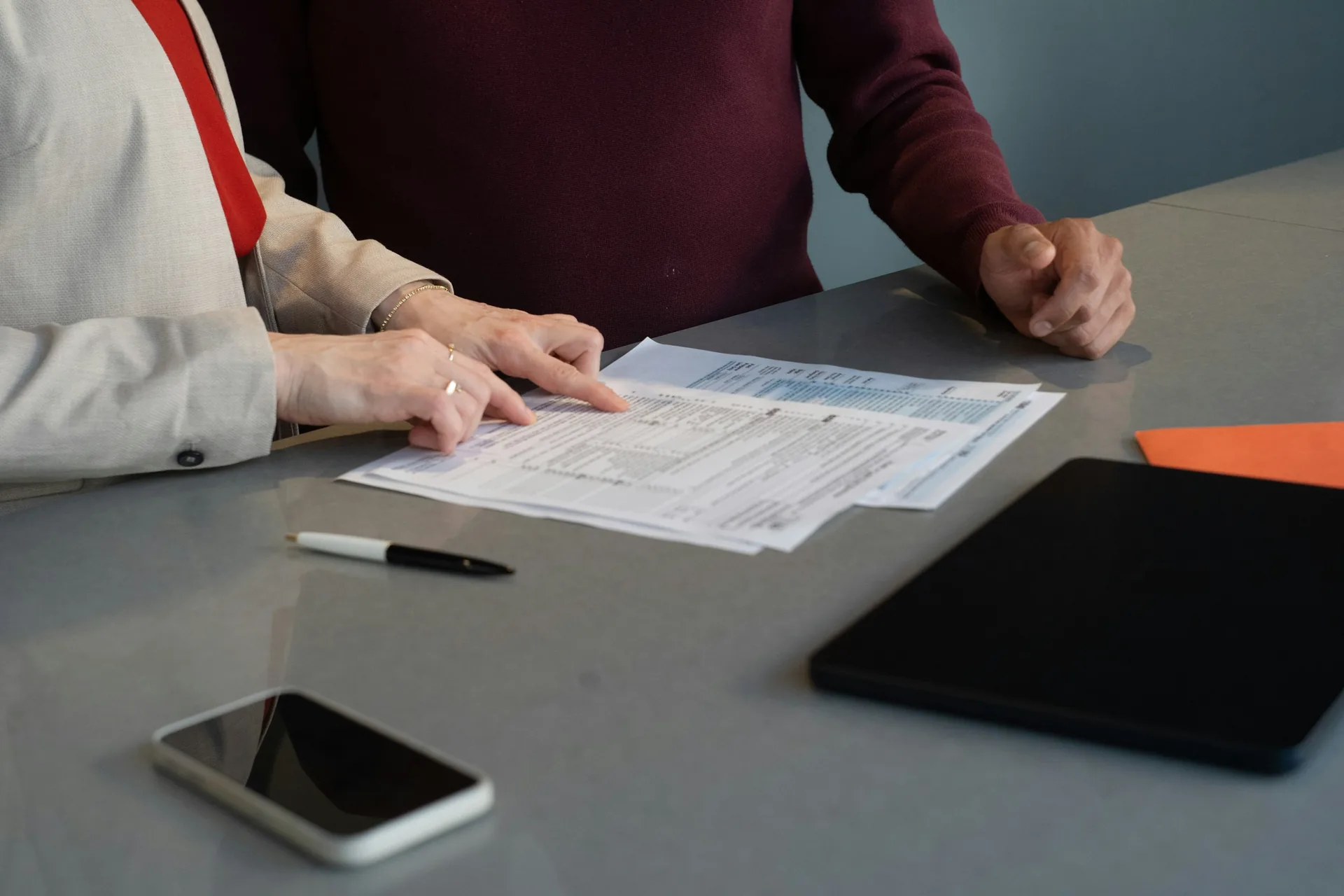 Two people reviewing a tax form on a gray desk, with a smartphone, tablet, and pen nearby