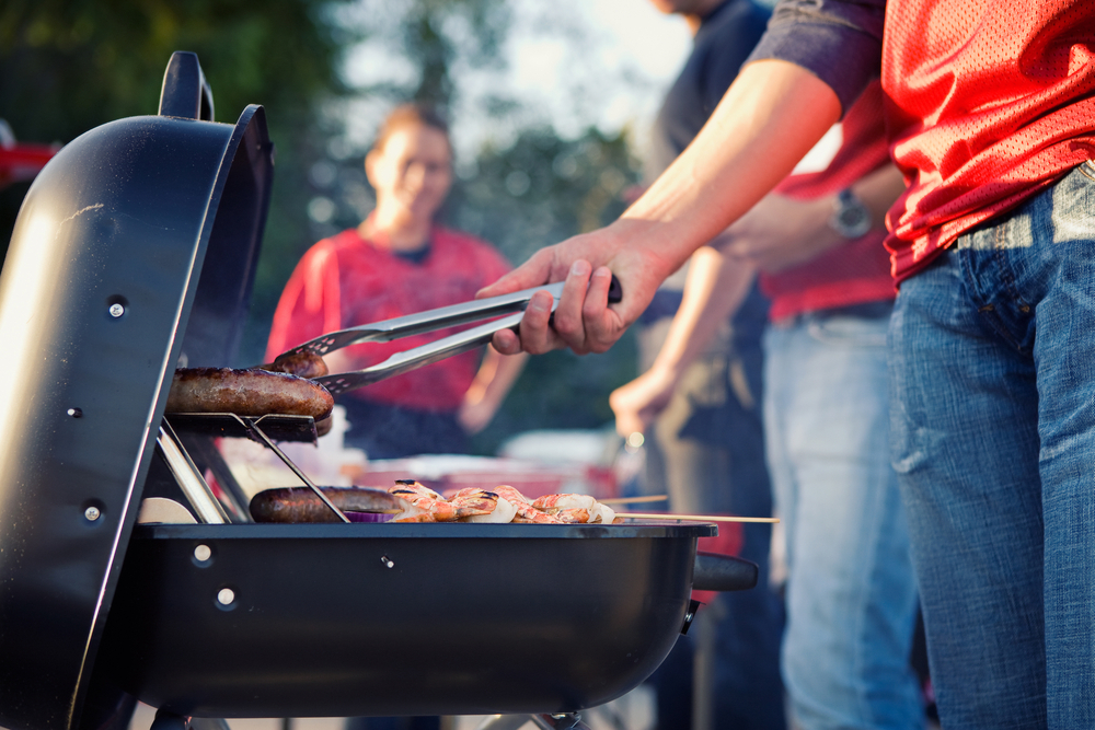 In focus is a grill cooking various meats, blurred in the background are 3 people hanging out all wearing the color red