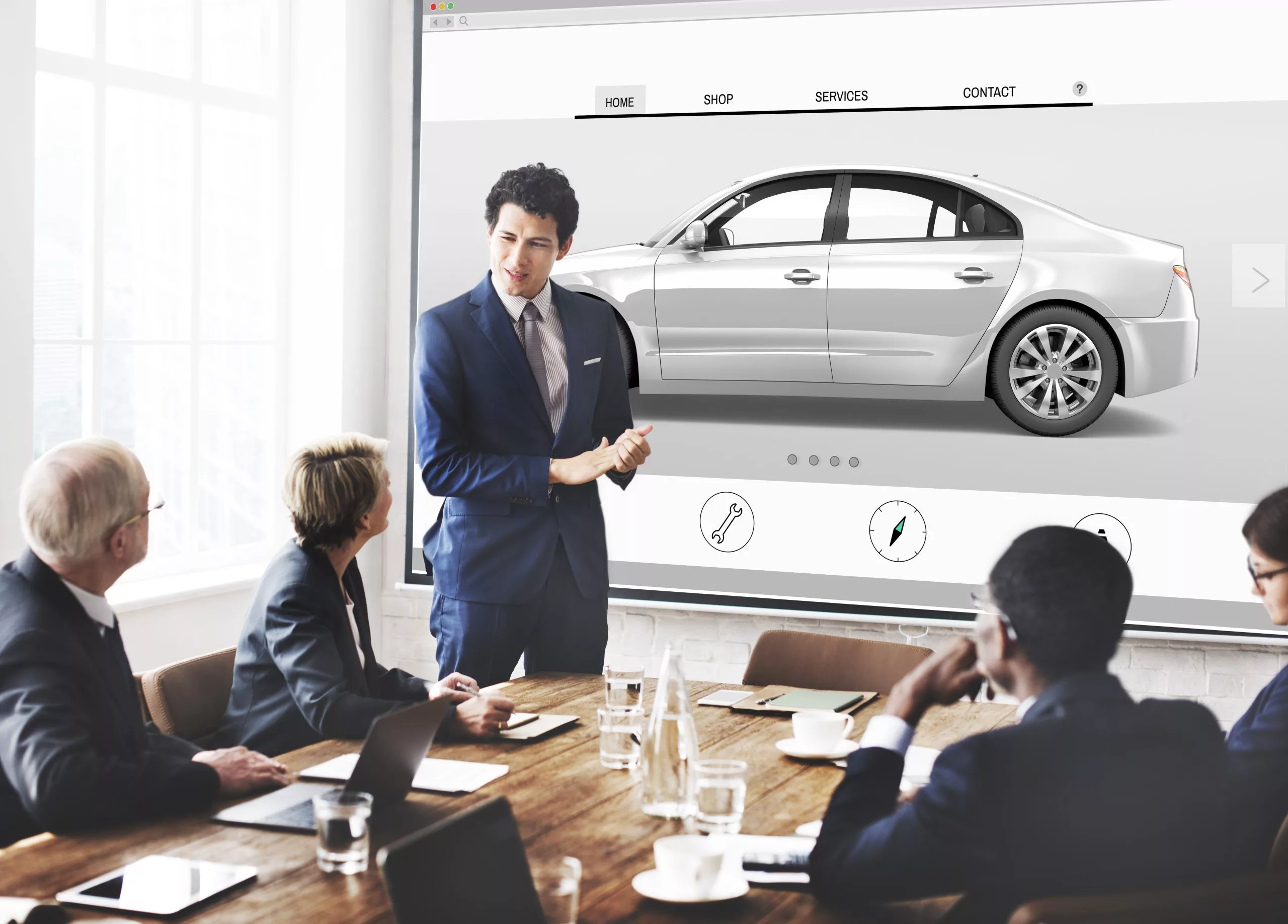 Boardroom with 4 people watching  another person  present a silver car a man is presenting with 2 females and 2 males watching on, everyone is in formal attire, on the presentation are 4 tabs, home, shop, services, and contact.