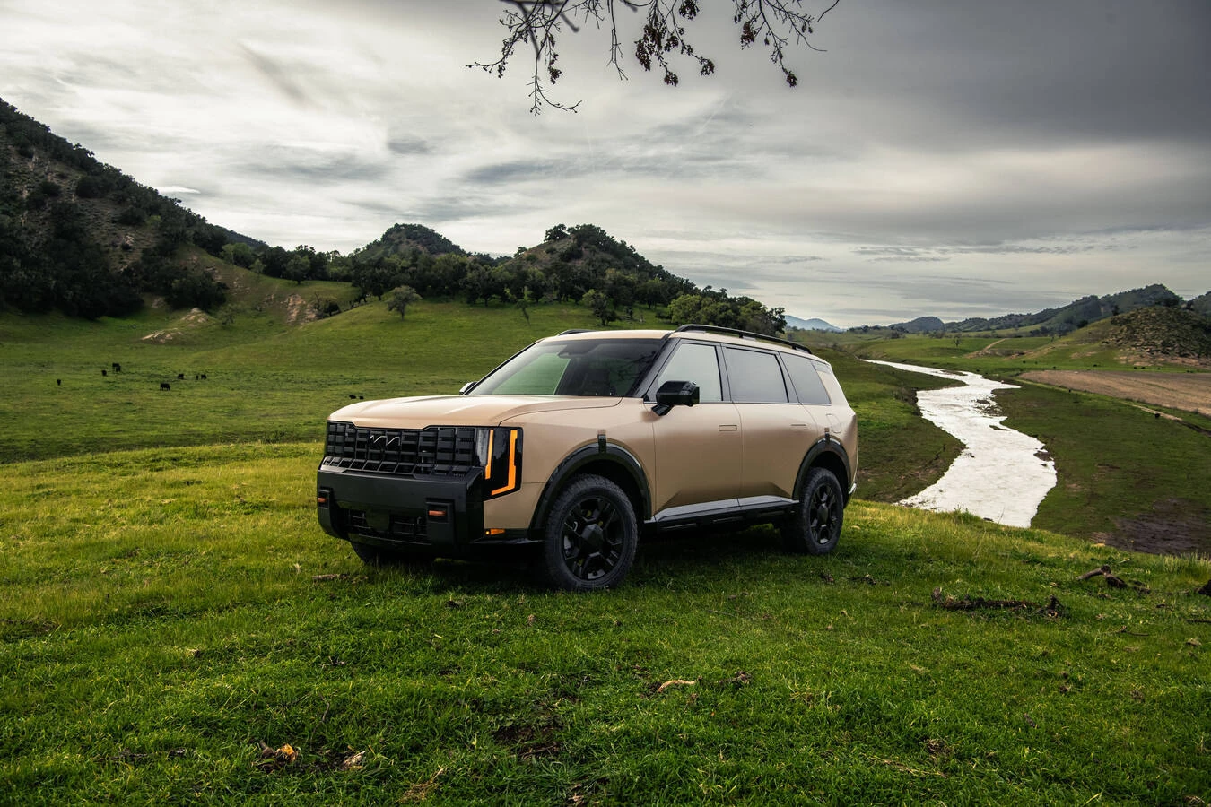 Matte tan 2027 Kia Telluride X-Pro parked on a grassy hillside above a winding river, with rolling hills, grazing cattle, and low clouds in the distance.