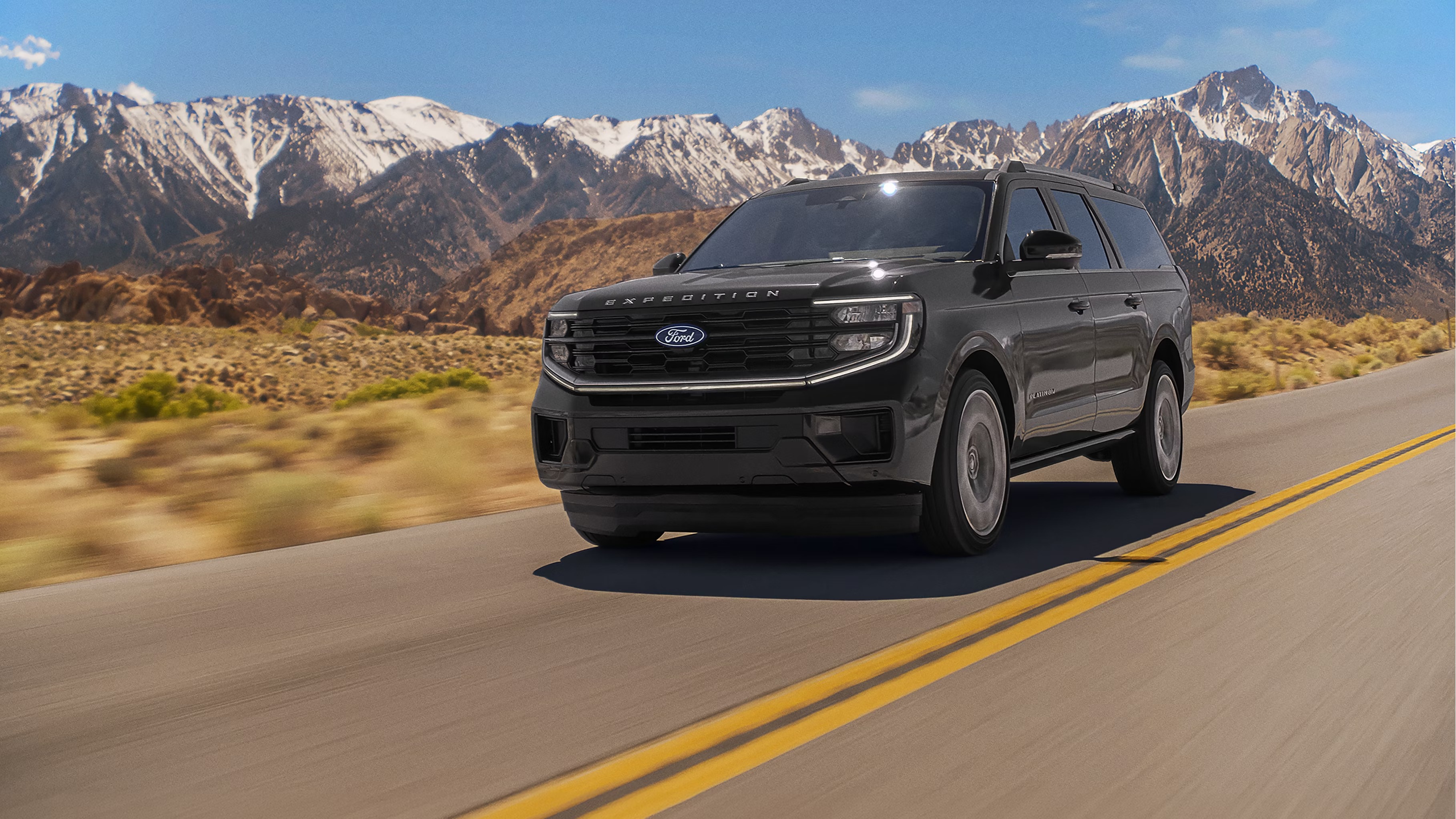 Black 2027 Ford Expedition Platinum SUV driving along a desert highway with snow-capped mountains in the background, shown from a front three-quarter angle with motion-blurred surroundings.