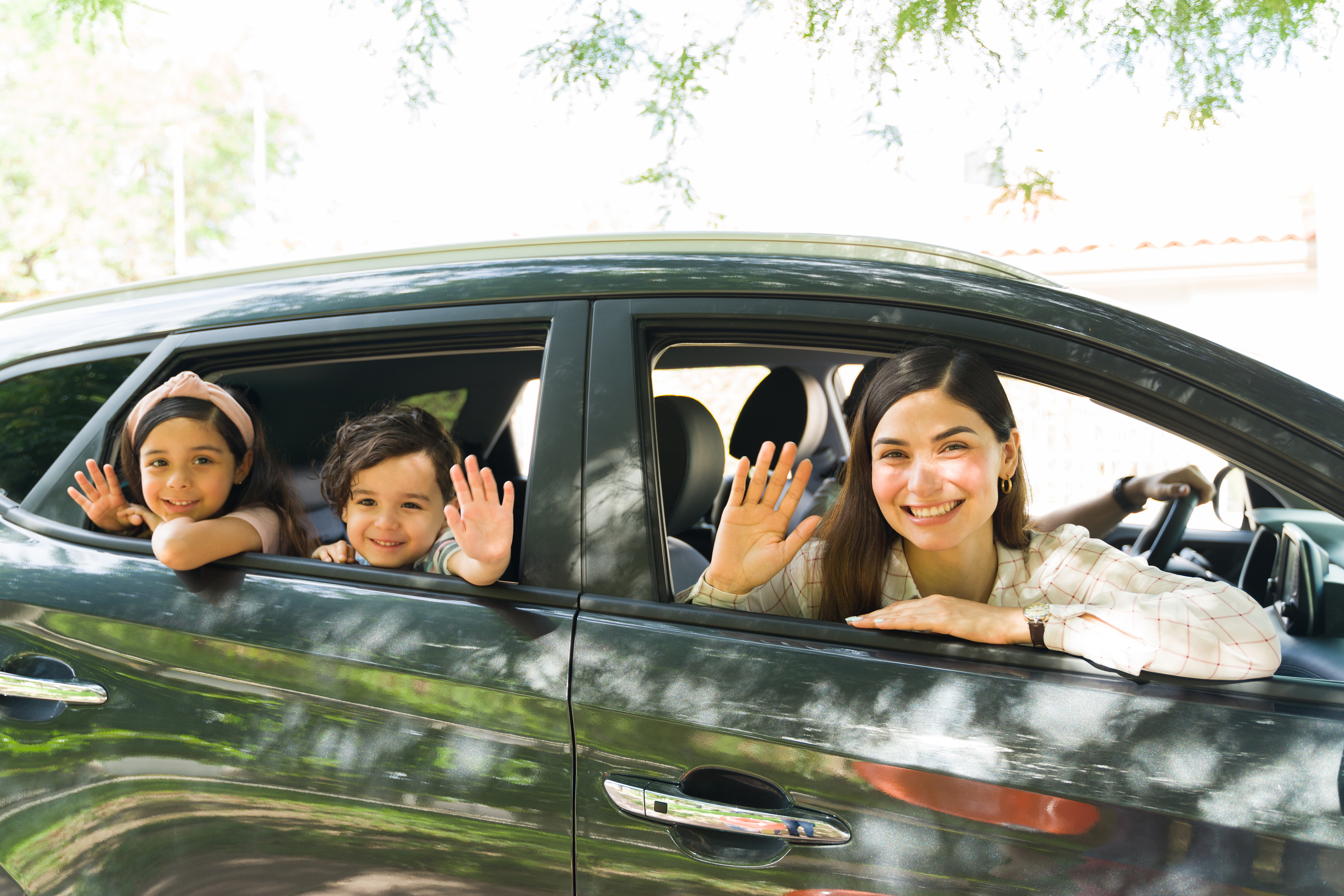 See you after the holidays. Beautiful mom and cute little brother and sister waving goodbye while sitting inside the car before a family trip