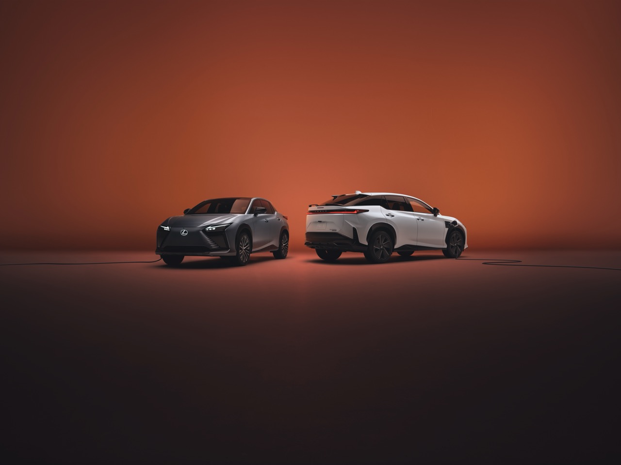 Two Lexus RZ electric SUVs, silver and white, plugged into chargers against a warm orange gradient background.