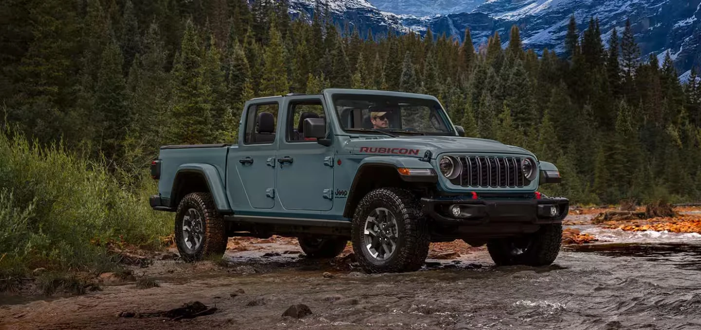 Blue-gray Jeep Gladiator Rubicon pickup with its top off, black fender flares and large off-road tires, crossing a shallow rocky stream with a driver at the wheel, evergreen trees and snow-capped mountains behind it