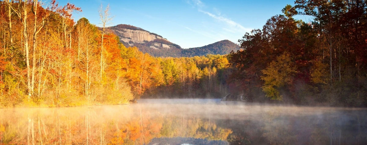 Autumn trees in orange, gold, and red lining a misty lake at sunrise, with a granite-faced mountain under a blue sky and the foliage reflected in the water.