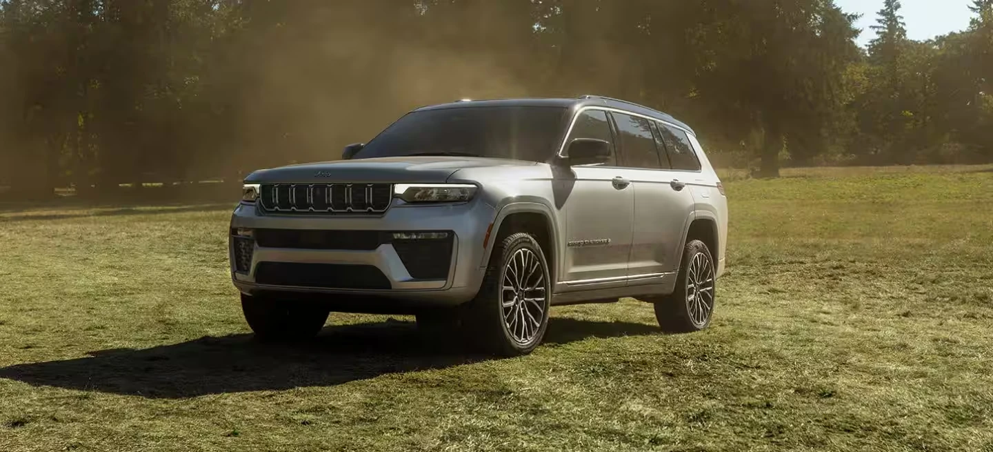 Silver 2026 Jeep Grand Cherokee L three-row SUV parked in a sunlit grass field with dust and trees behind it