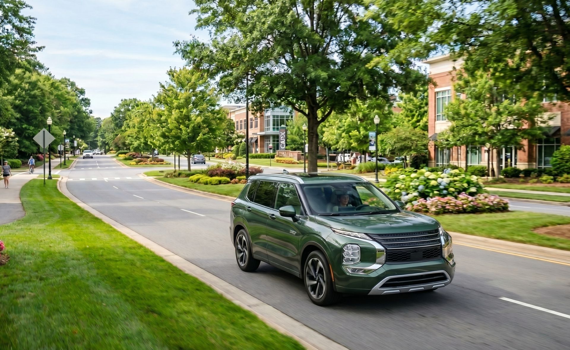A metallic green Mitsubishi Outlander SUV driving down a scenic, tree-lined suburban boulevard past modern brick buildings and manicured green lawns.