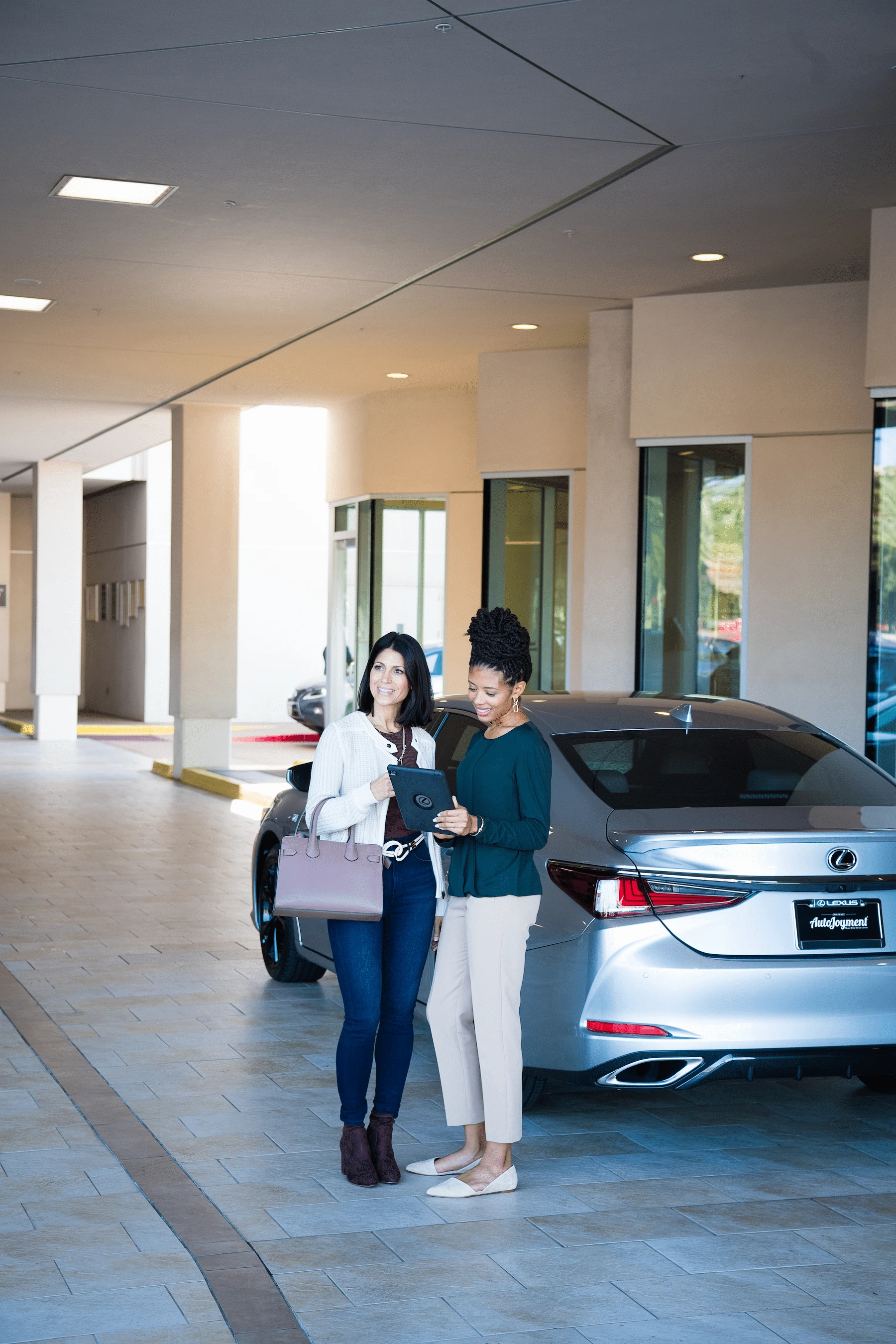 A Lexus sales associate and customer reviewing financing options on a tablet beside a silver Lexus sedan under a dealership breezeway.