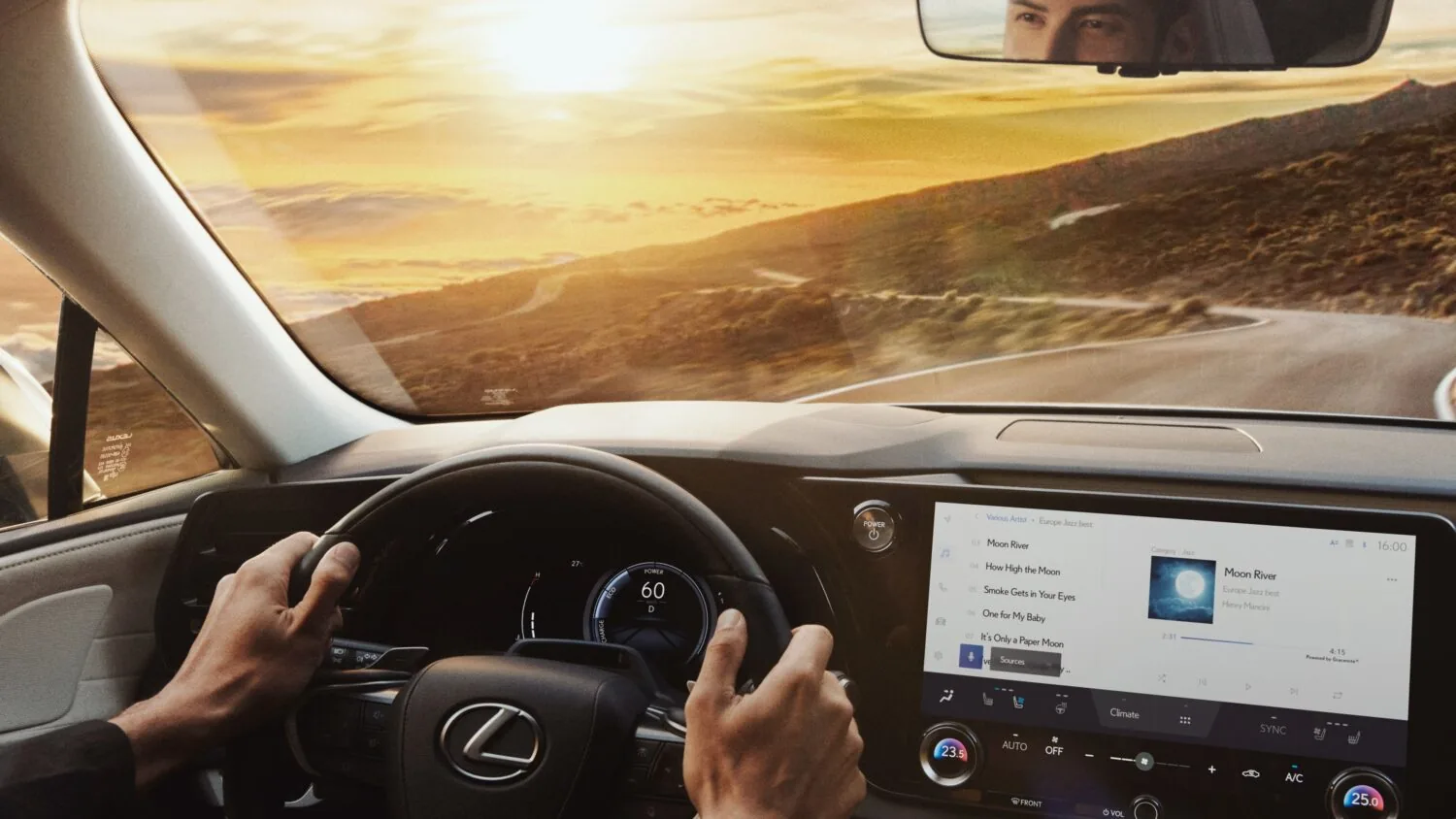 Point-of-view from the driver's seat of a Lexus showing hands on the steering wheel with the Lexus logo, a digital speedometer reading 60 mph, and an infotainment screen displaying a music playlist, with a sunset over a winding road ahead.