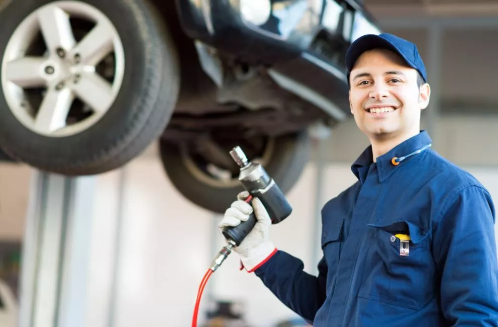 Bottom of Car is up on forklift, Mechanic stands closer to frame smiling wearing a blue jumpsuit holding a tool