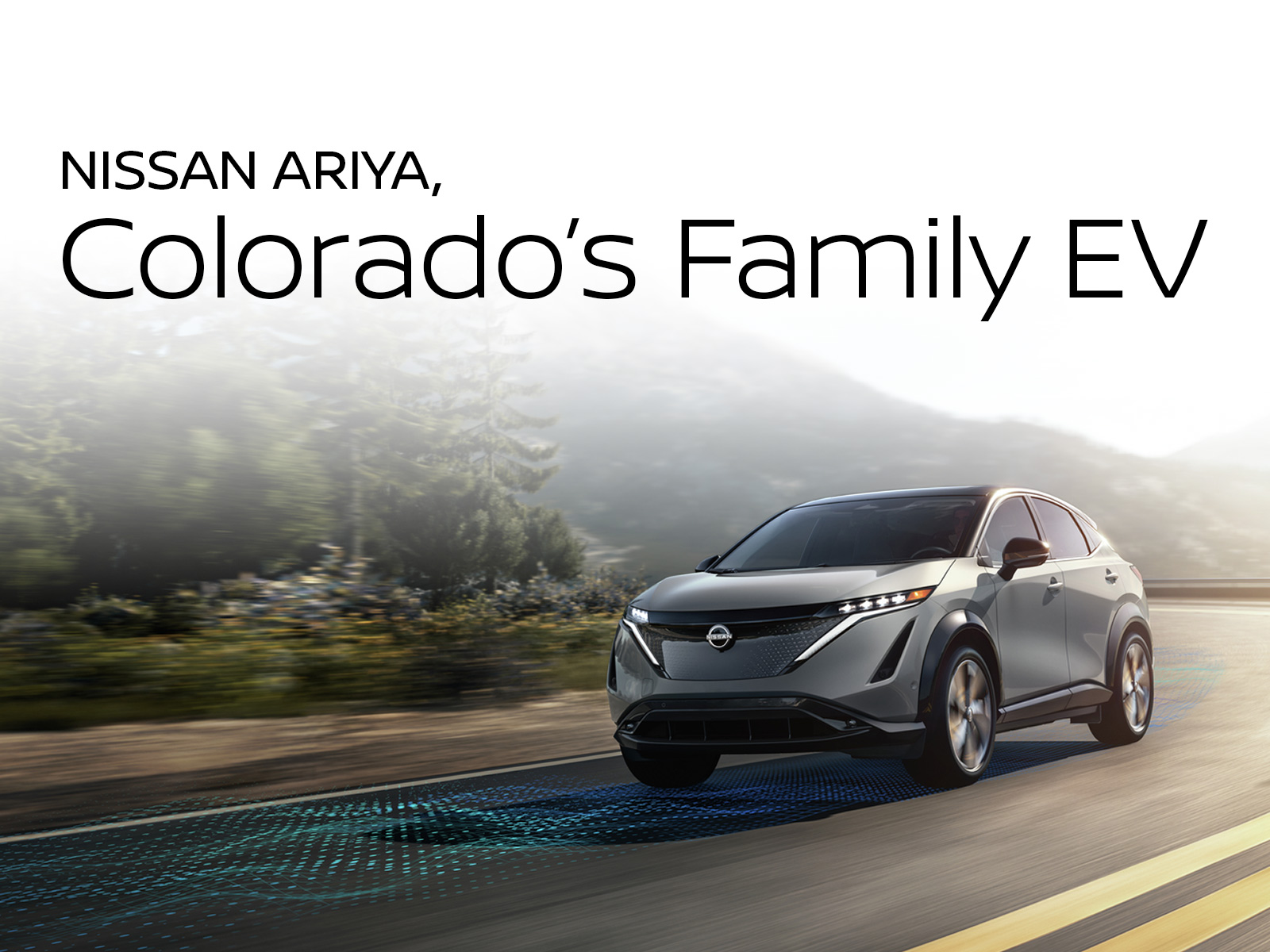Nissan ARIYA electric SUV driving on a scenic Colorado road with mountains and forest in the background. Text overlay says “Nissan ARIYA, Colorado’s Family EV.