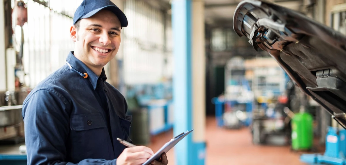Smiling technician in a navy cap and coveralls holding a pen and clipboard in a bright service garage, beside a vehicle with its hood raised.