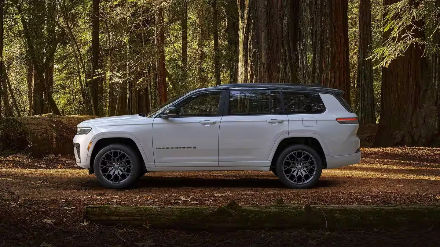 White 2027 Jeep Grand Cherokee L three-row SUV with a black roof and dark multi-spoke wheels, shown in side profile in a forest clearing among tall redwood trees with fallen logs