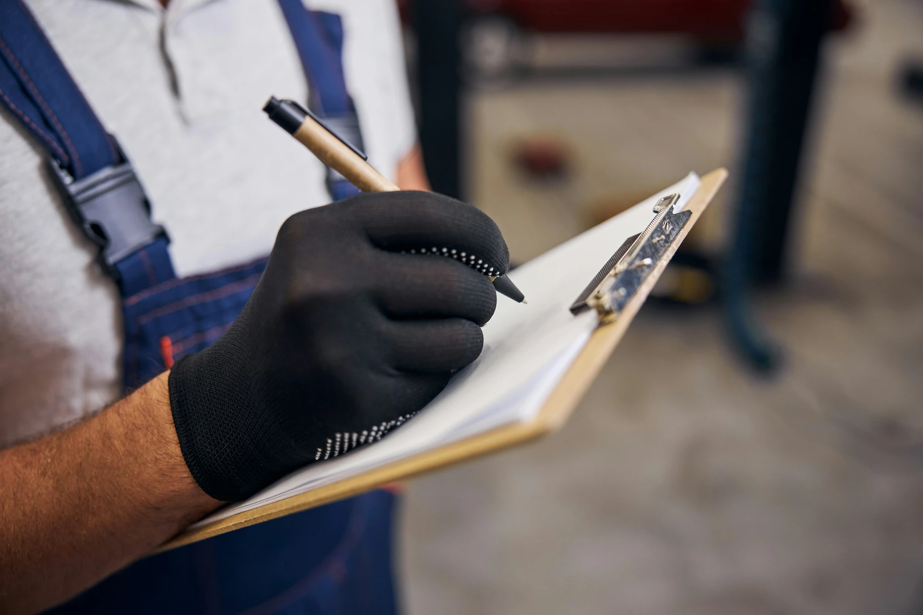 Gloved hand writing on a clipboard checklist, referencing a technician documenting a vehicle recall inspection, with a blurred workshop background