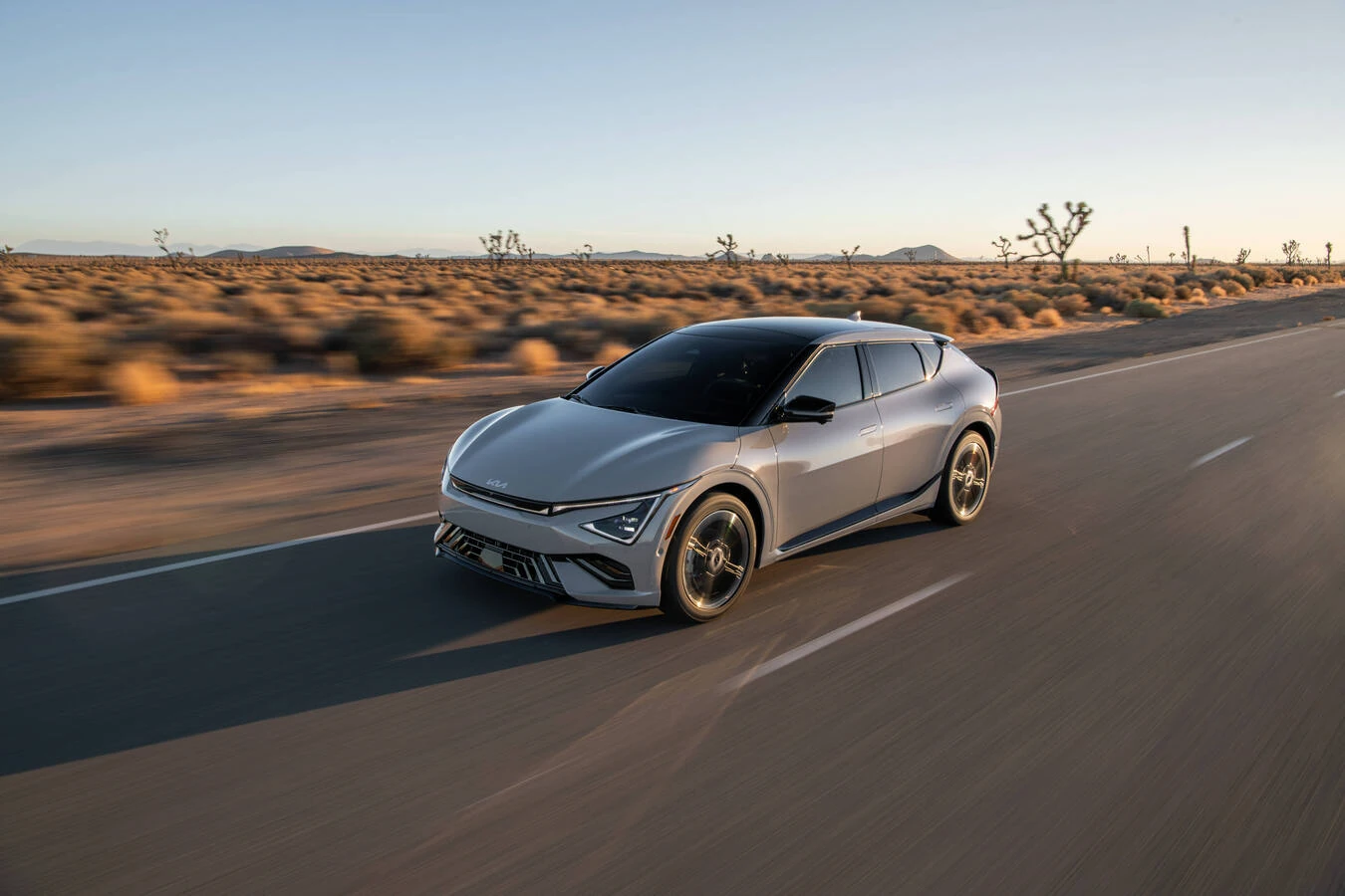 White Kia EV6 driving on a two-lane desert highway at dusk, with Joshua trees and mountains in the background