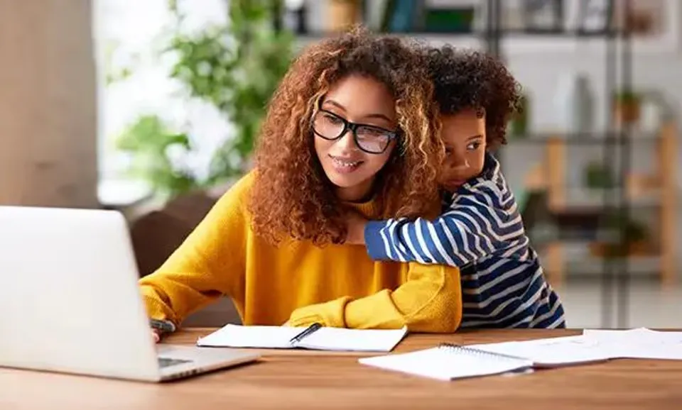 Trading in a vehicle when purchasing can lower your monthly payments and save money on taxes. Smiling mother wearing cozy yellow sweater is researching trade values on computer and taking notes with pen and paper. Her son hugging her while she works.
