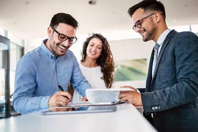 On the left side of a table are two people, a man and a woman, the man is standing with a smile signing a piece of paper, the woman stands behind him also smiling, on the right side of the table stands a man who is giving the man the paper to sign, out of focus in the background is a painting and the table is white