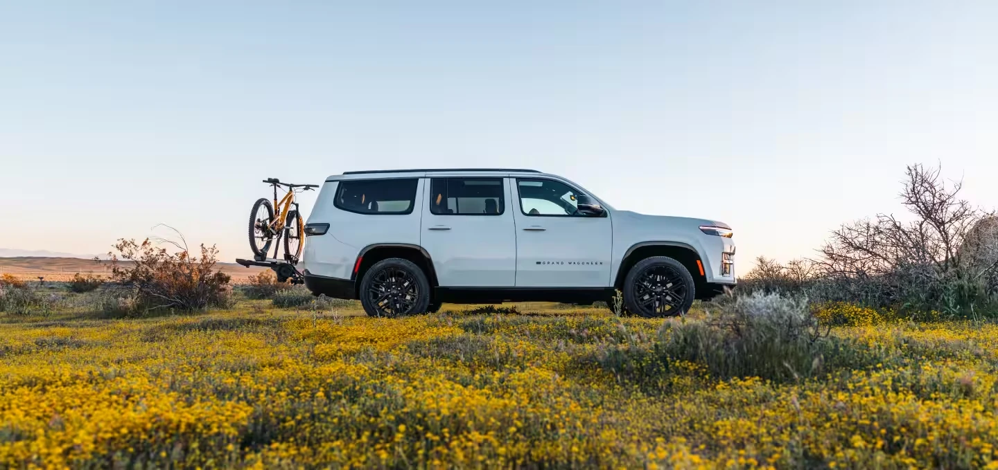 White 2027 Jeep Grand Wagoneer SUV with black wheels and a hitch-mounted rack carrying a yellow mountain bike, in side profile in a field of yellow wildflowers with desert shrubs under a pale sky