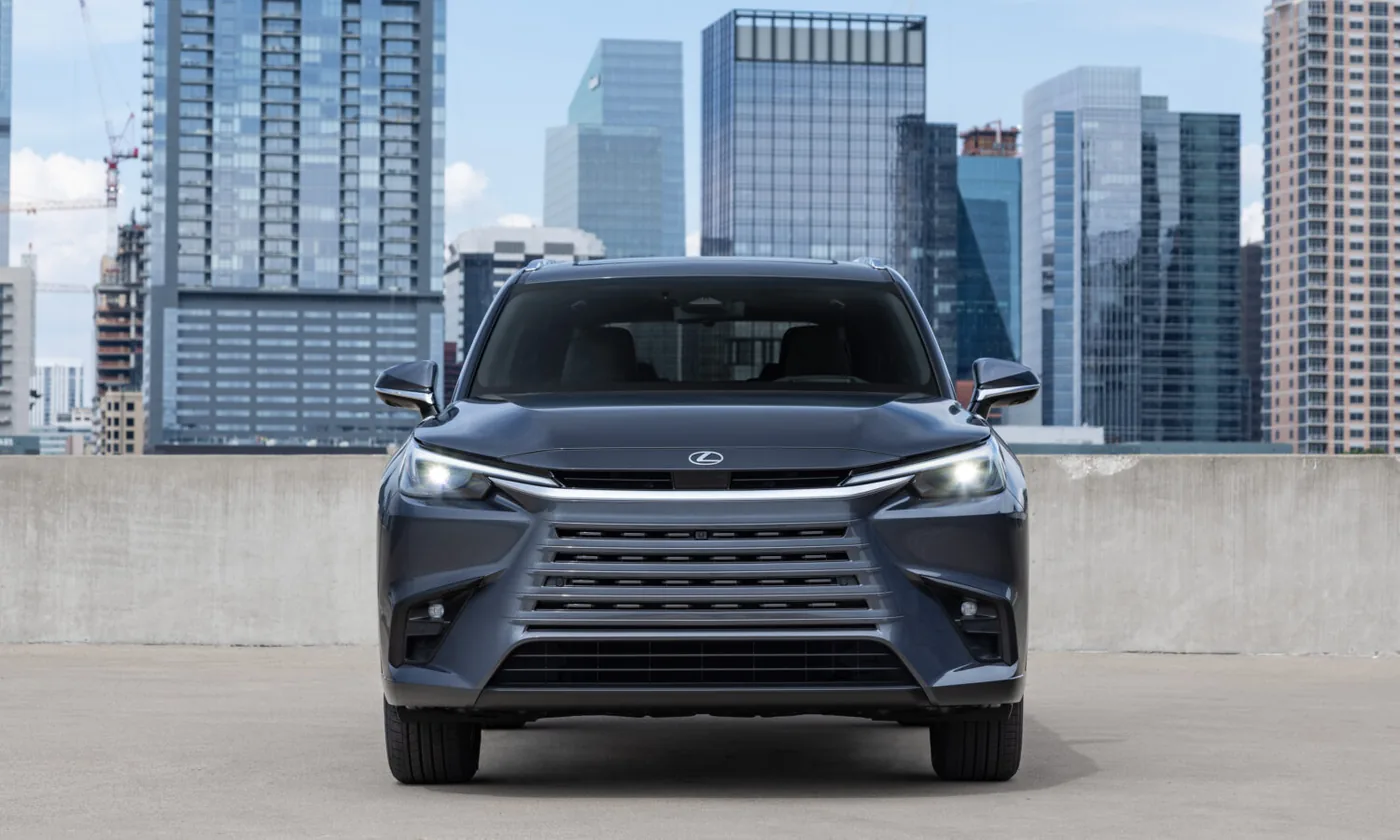 Head-on view of a dark gray Lexus SUV with a horizontal-slat grille and LED headlights, parked on a concrete rooftop deck in front of glass high-rise buildings under a partly cloudy sky.