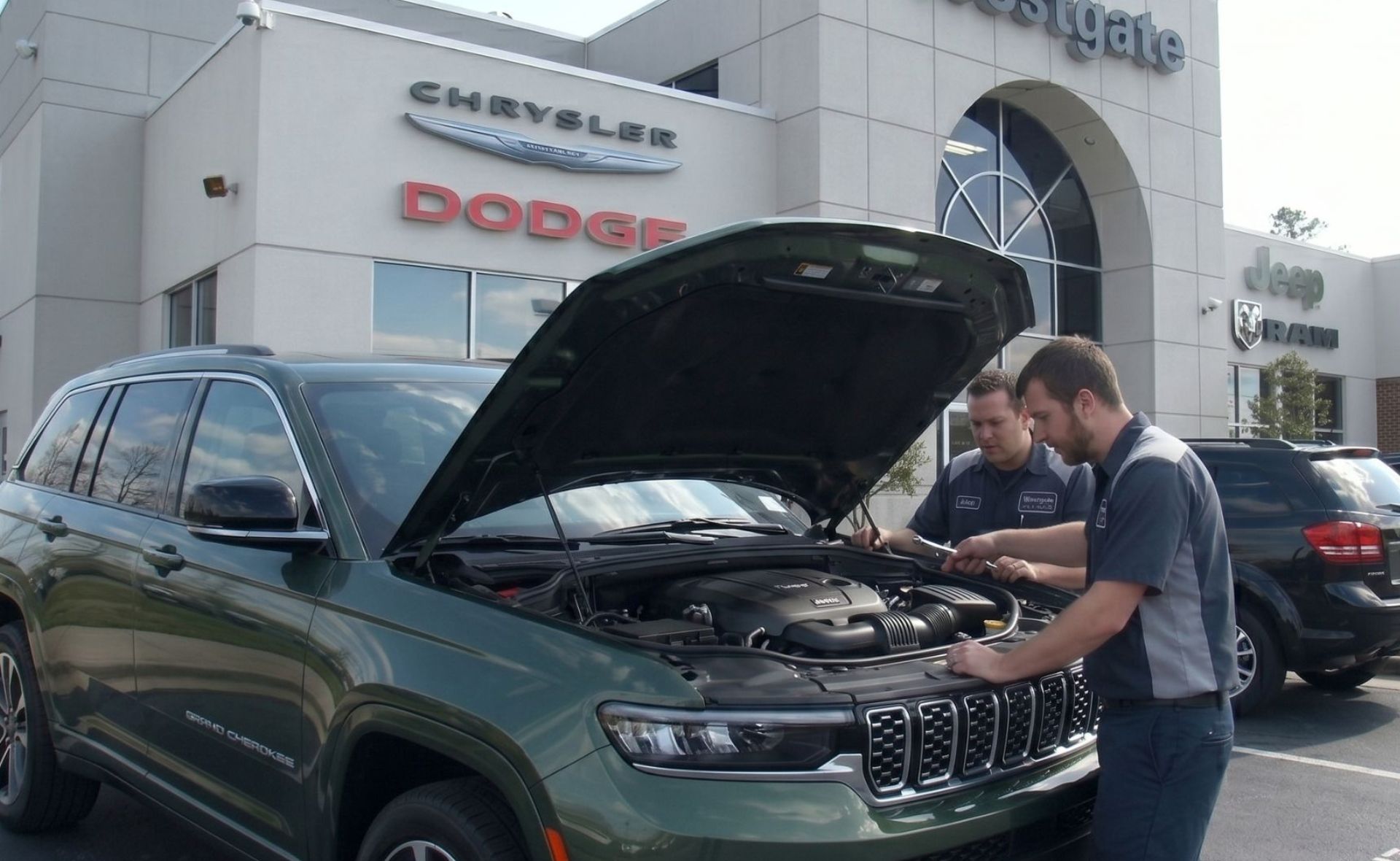 Two automotive service technicians inspecting the engine bay of a green Jeep Grand Cherokee SUV with its hood open in the lot outside the Westgate Chrysler Jeep Dodge Ram dealership in Raleigh.