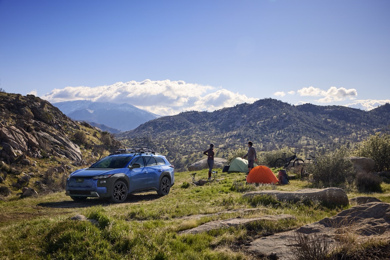 A static image of a blue 2026 Subaru Trailseeker parked on a grassy clearing in a rocky mountain landscape. 