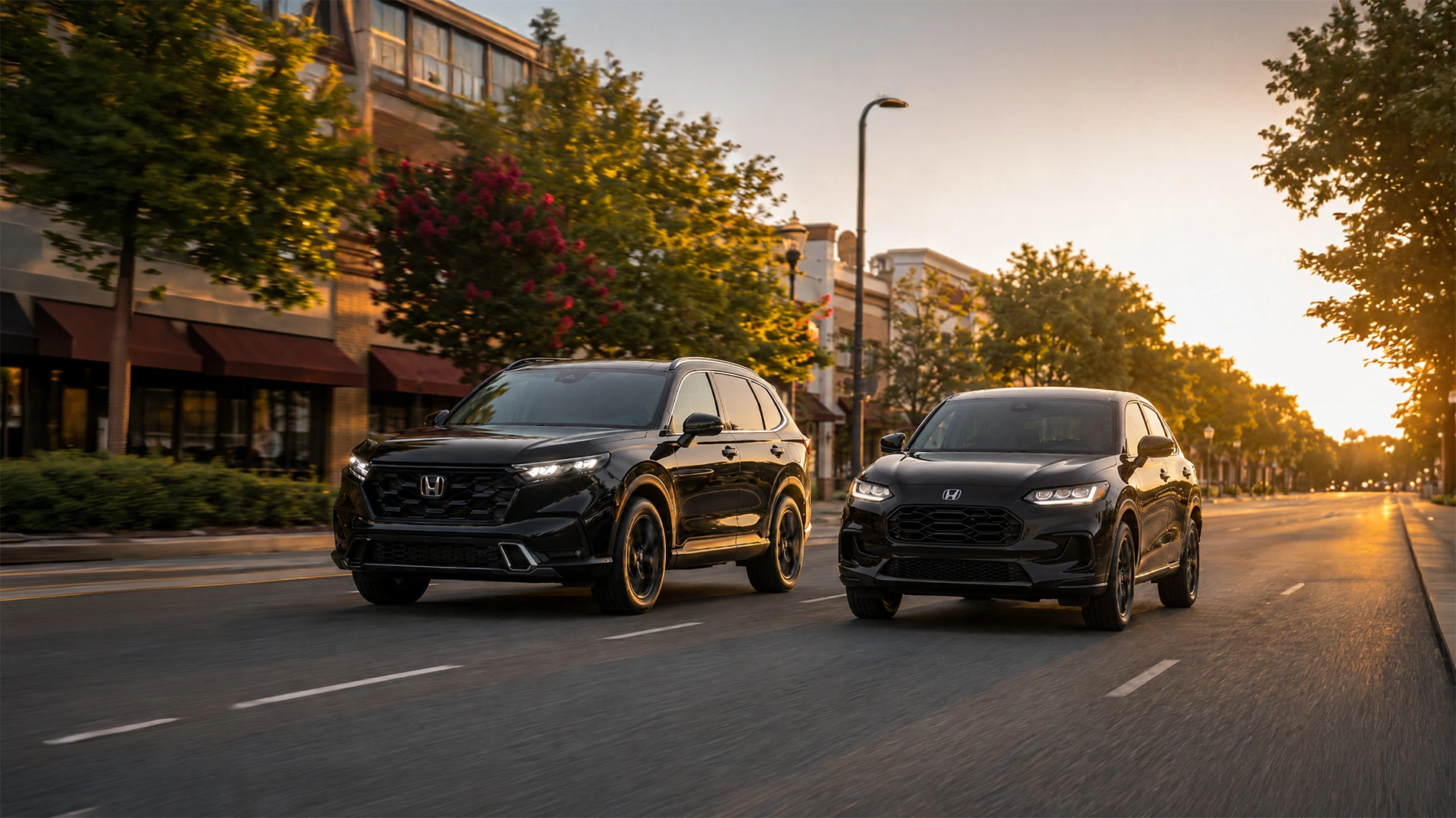 Black Honda CR-V and Honda HR-V driving through a downtown street at sunset with modern city buildings and tree-lined roads in the background.