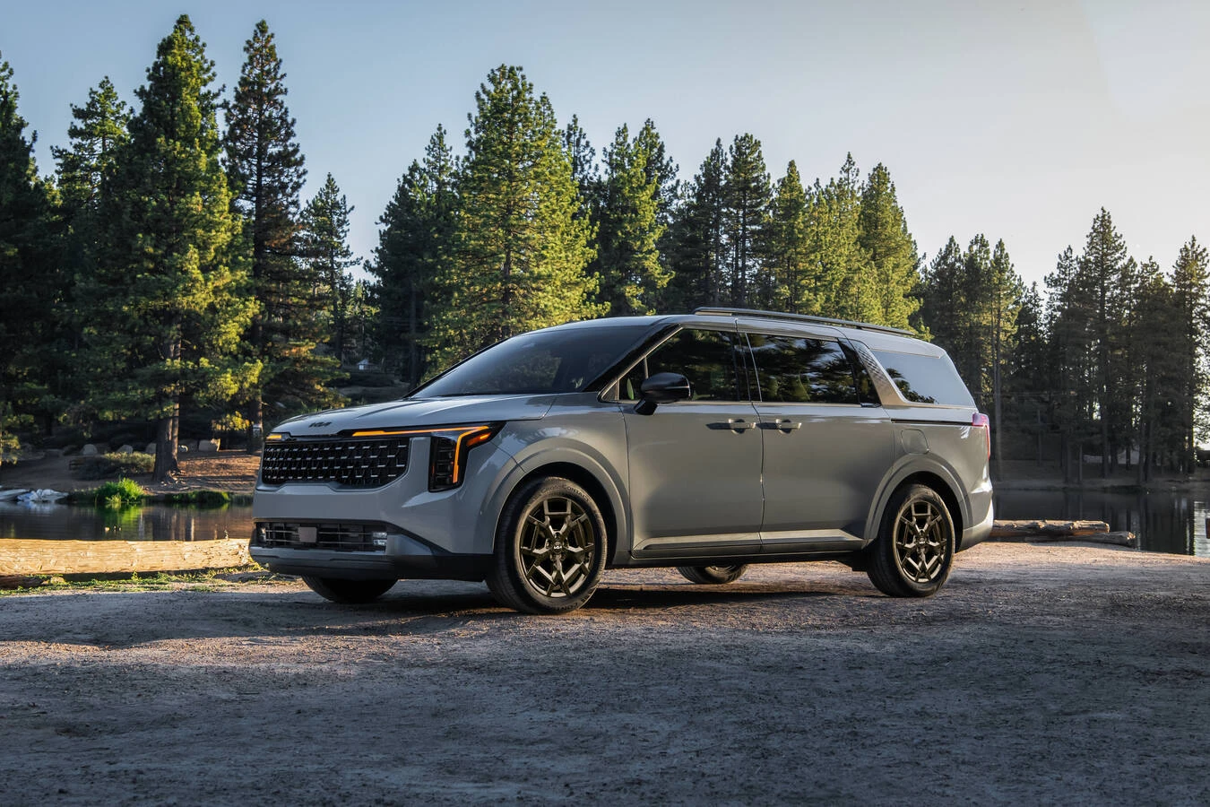 Silver 2027 Kia Carnival Hybrid minivan parked beside a lake with pine trees and a wooden dock along the shoreline.