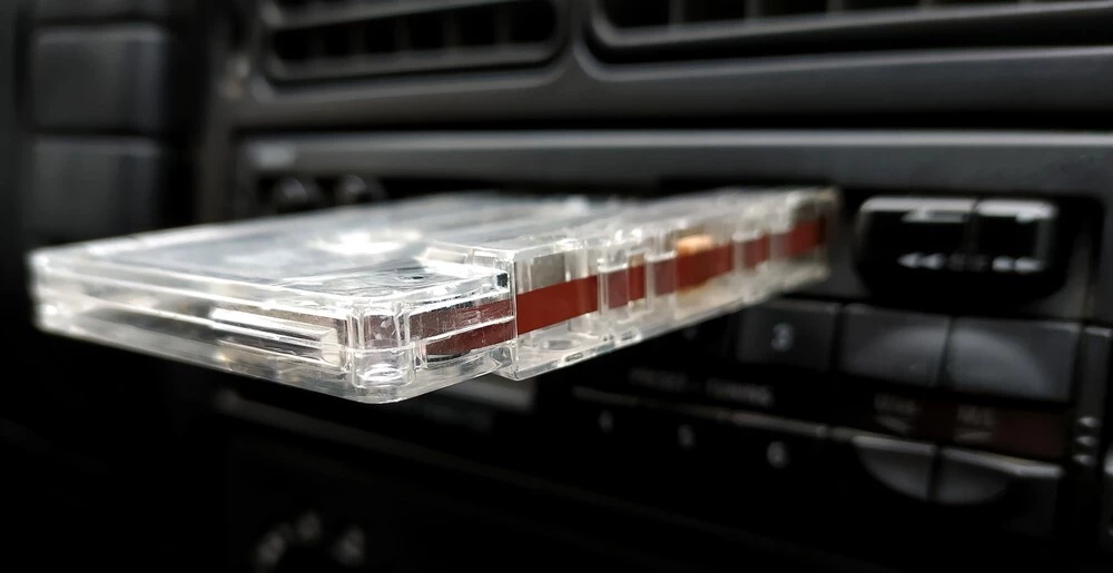 A Cassette is focused in on and is inserted into a cars Cassette player, the cars interior is in the background blurred out and black