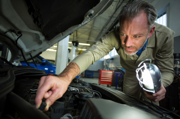 A mechanic leans into an open hood of a car, shining a work lamp into the engine bay.