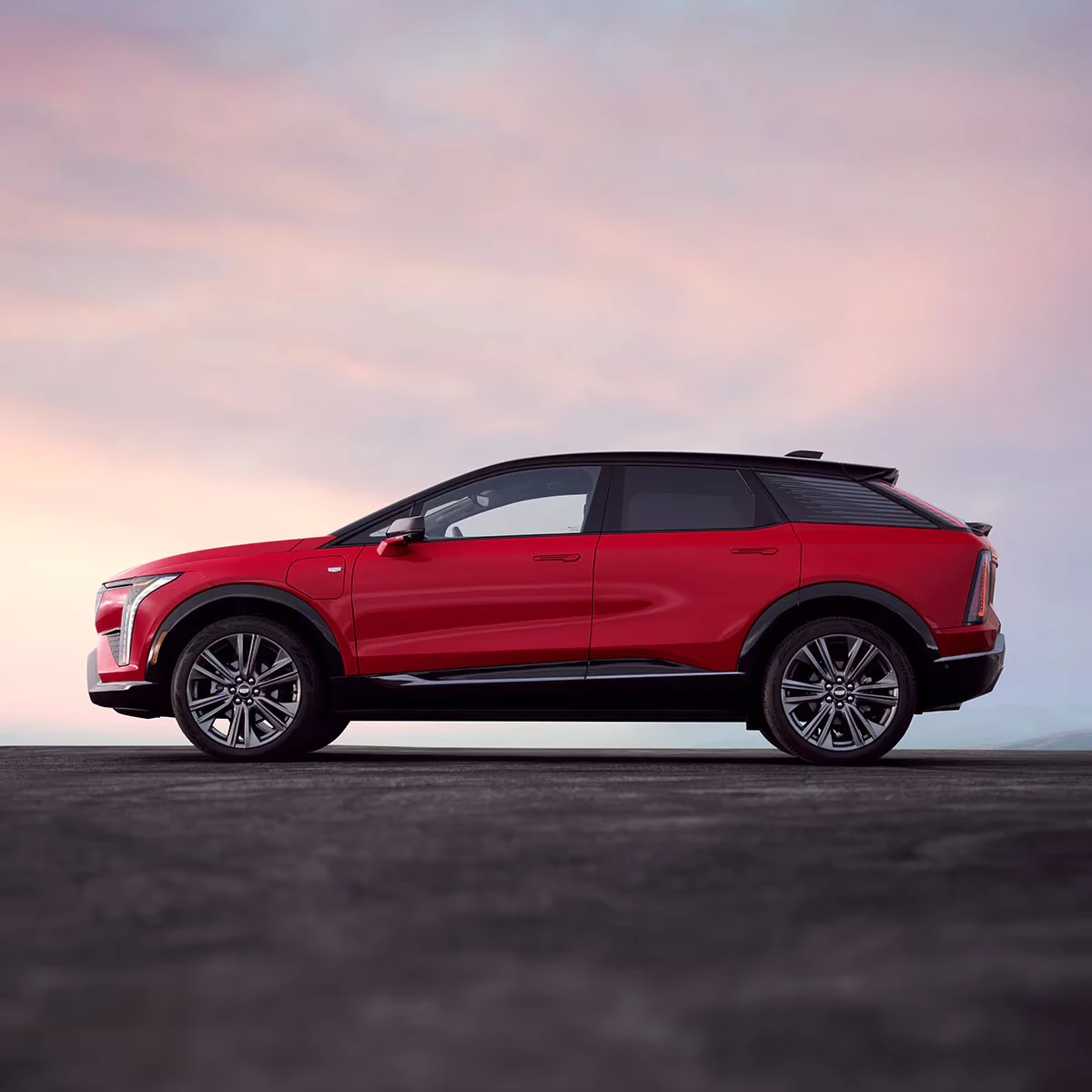 Red Cadillac Optiq SUV in side profile with a black roof, rear roof spoiler, black lower body trim and silver multi-spoke wheels, on a dark paved surface under a soft pink and lavender sky
