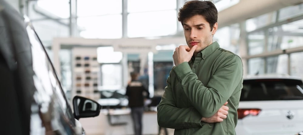 A man in a green polo is curiously studying a car in front of him, only the side mirror of the car in question is in frame, the setting is car dealership and blurred in the background is another car, a salesperson, and the rest of the dealership