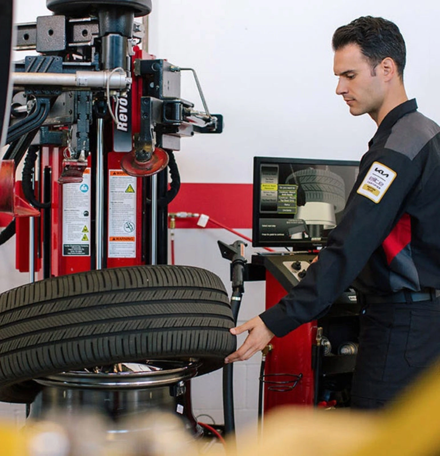 Kia Certified technician operating a tire balancing machine, checking a tire mounted on the machine's spindle with a diagnostic screen showing tread data in the background.