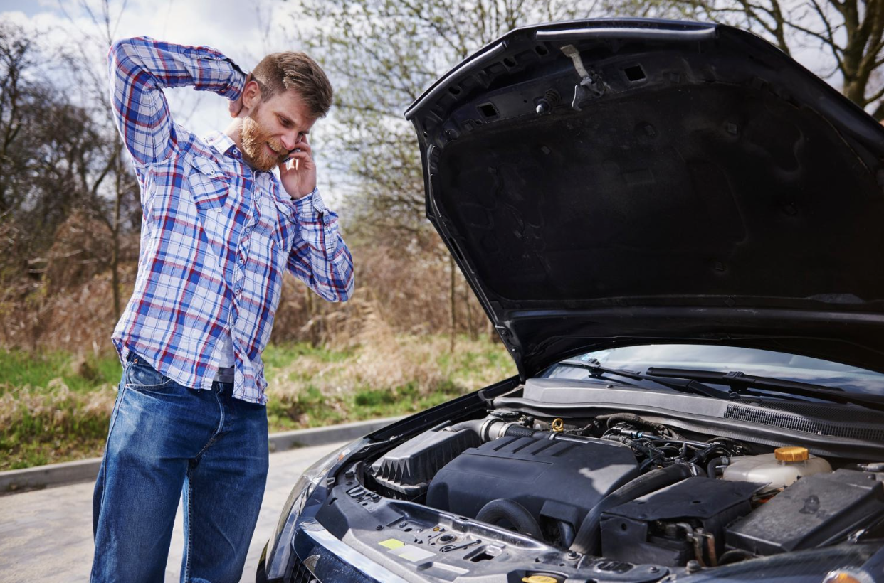 Frustrated man on the phone next to his broken-down car with the hood up.