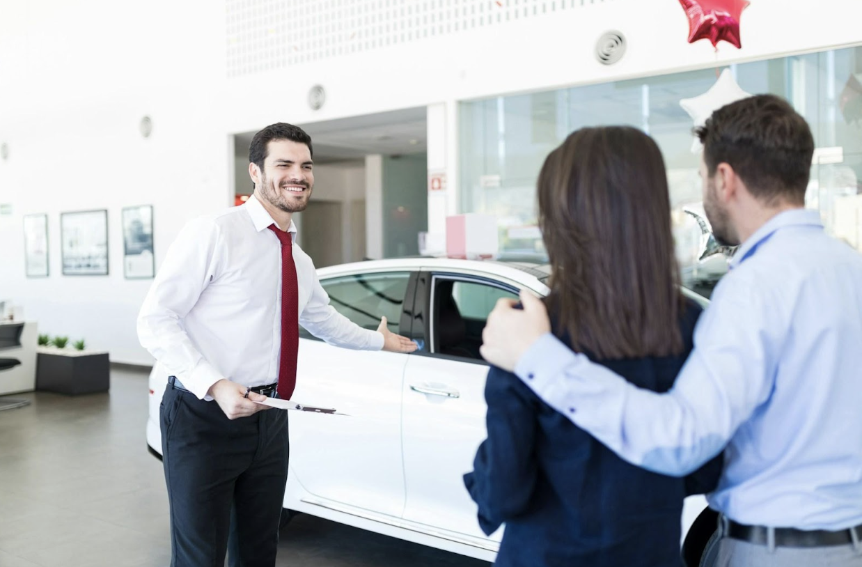 A smiling car salesman is gesturing toward a white car while talking to a couple in a dealership.