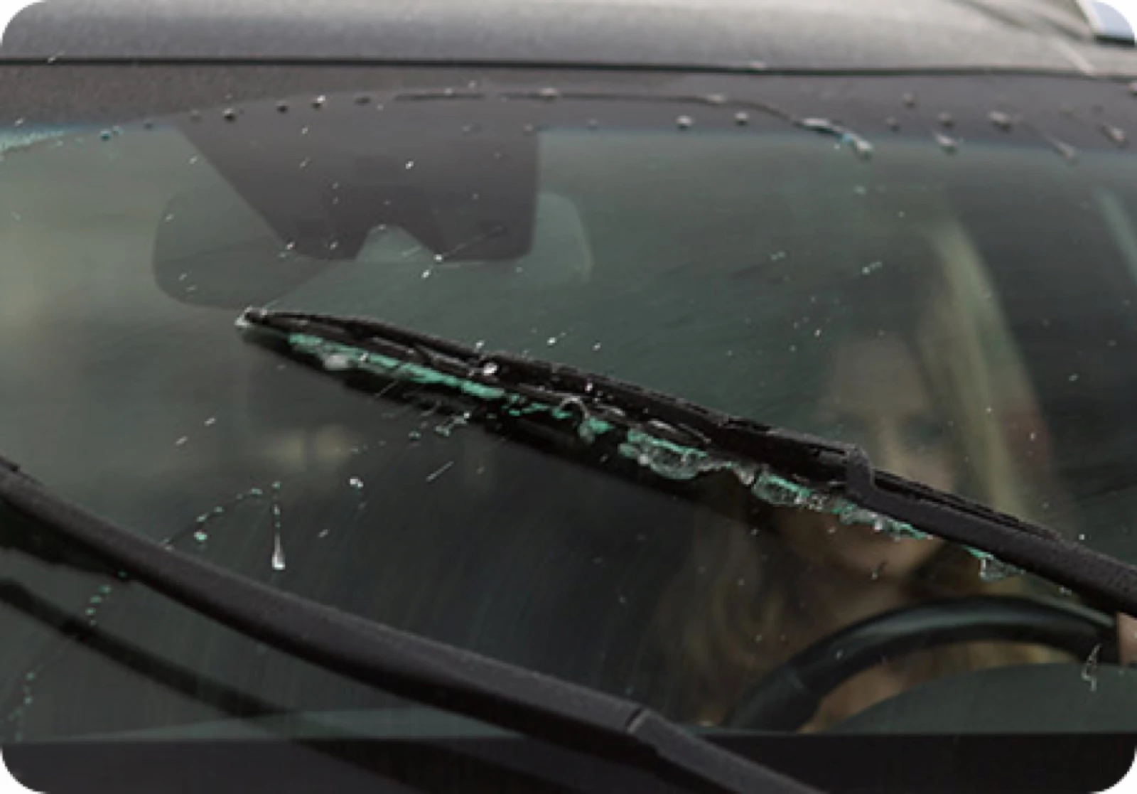 Close-up of a worn, cracking wiper blade on a wet windshield, with a driver visible blurred inside the vehicle.
