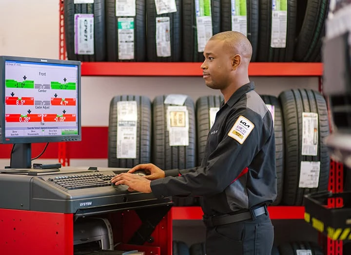 A Kia technician reviews front wheel alignment readings (camber, caster, and toe) on a diagnostic screen, with tire inventory shelved behind him.