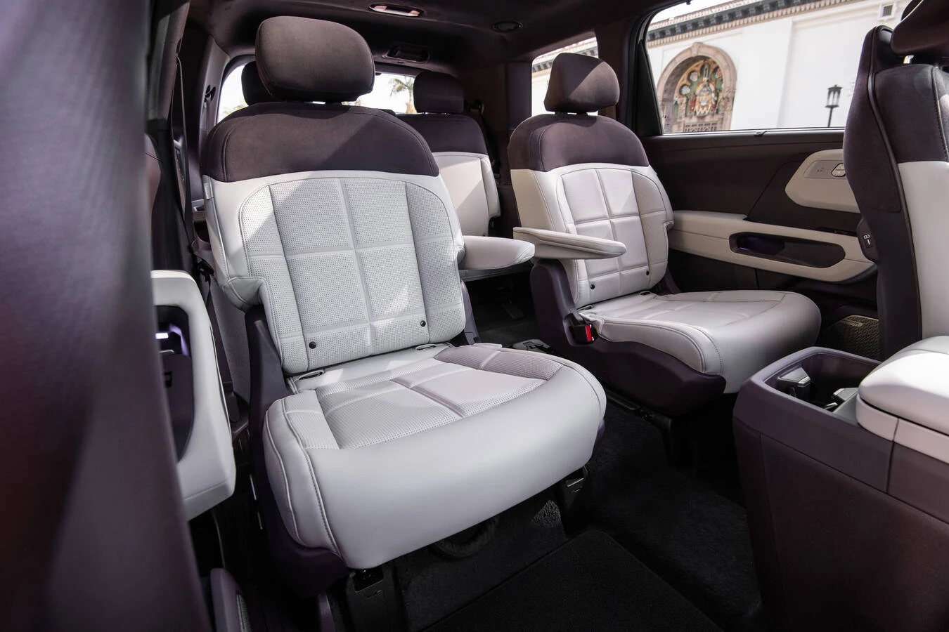 Second-row captain's chairs in a 2027 Kia Telluride HEV, cream leather with dark brown bolsters and headrests, viewed through an open door with an ornate building visible outside.