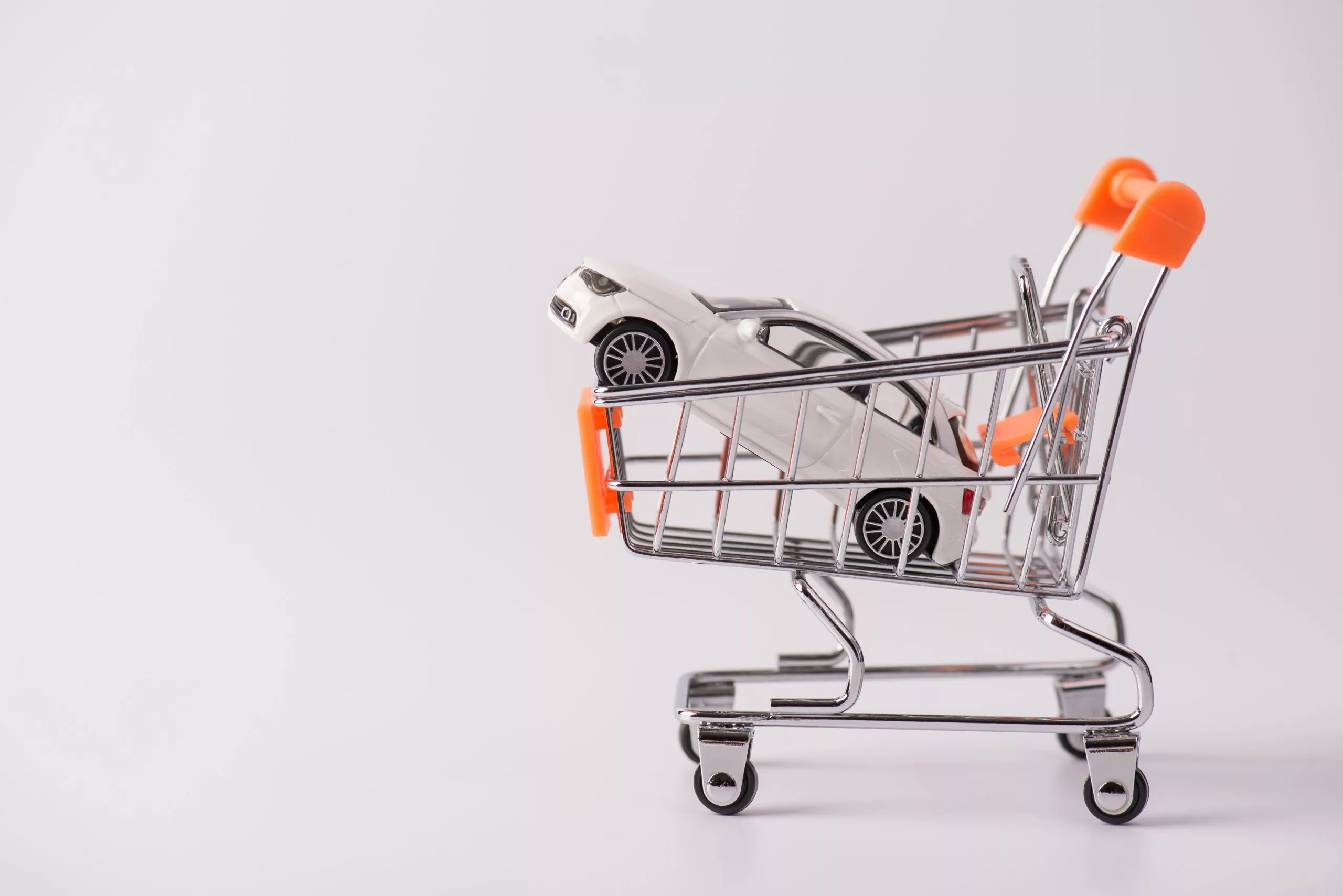 Miniature white toy car is in the basket of a shopping cart approximately a little larger than the toy car, the shopping cart has orange handles and the background is blank