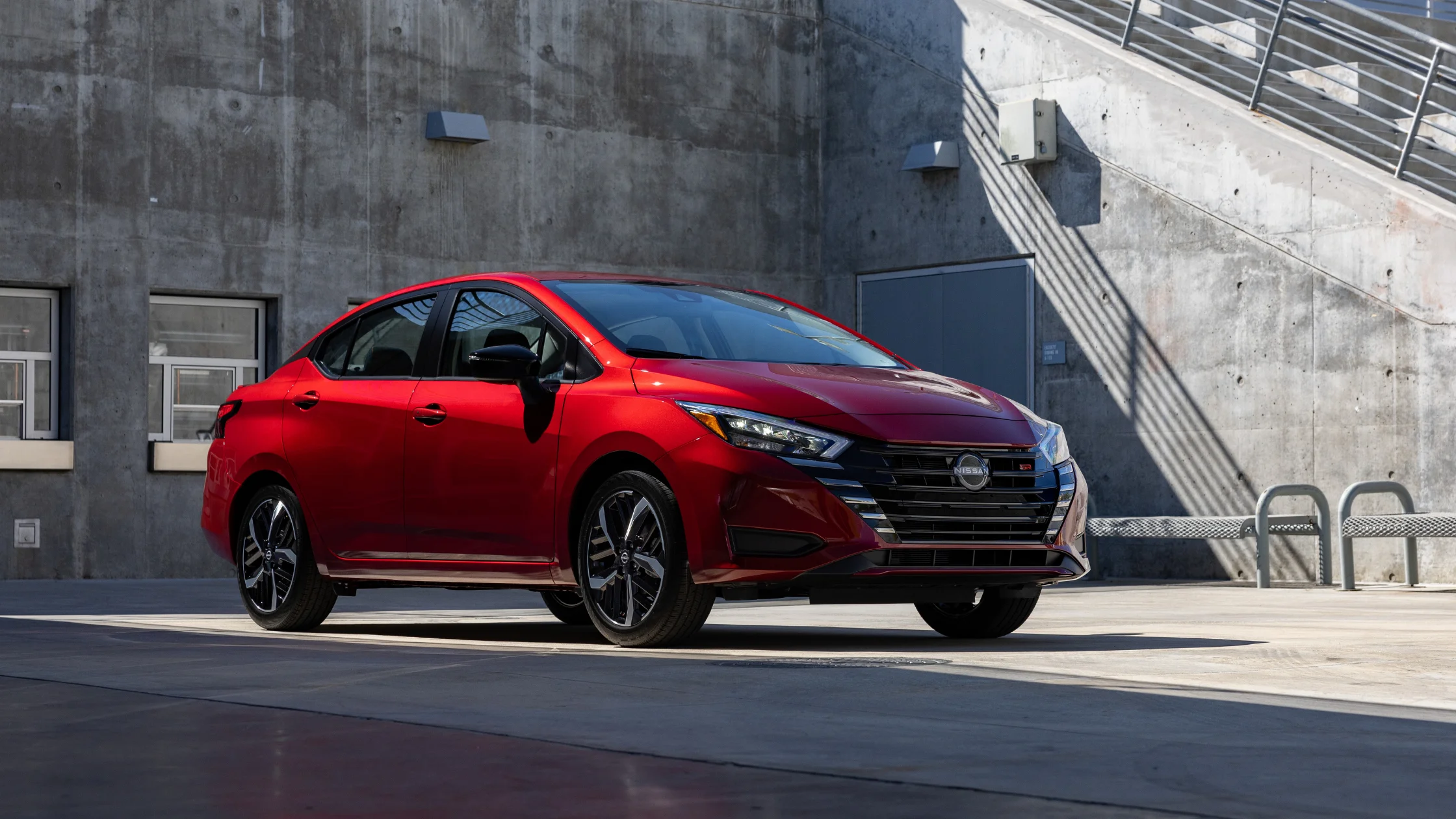 2025 Nissan Versa® red sedan parked by concrete building with black grille and sporty wheels