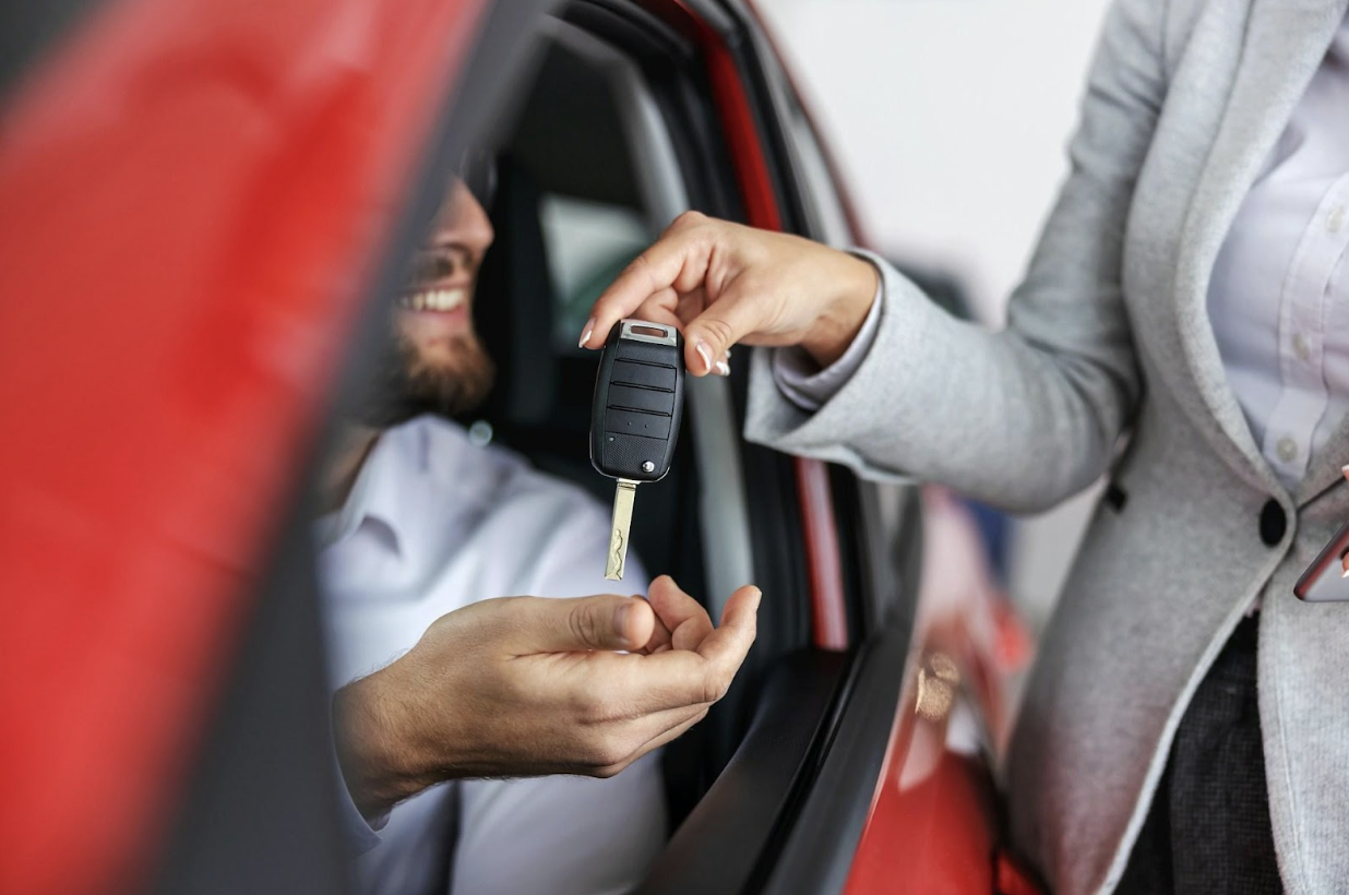 A salesperson handing the key to a new car to a smiling man in the driver's seat.