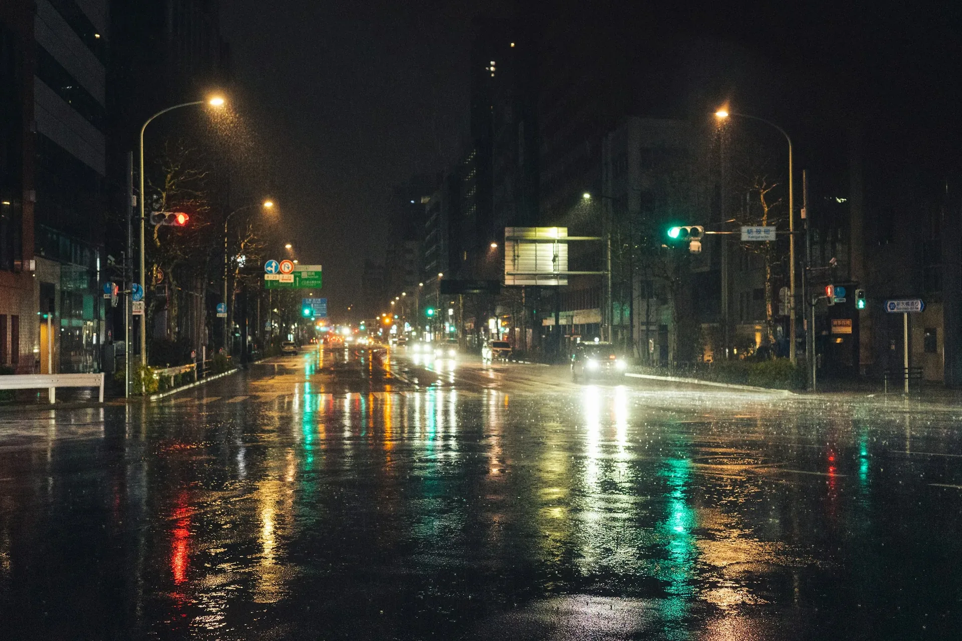 Wet city street at night with glowing headlights, traffic lights, and illuminated storefront signage reflecting off the pavement