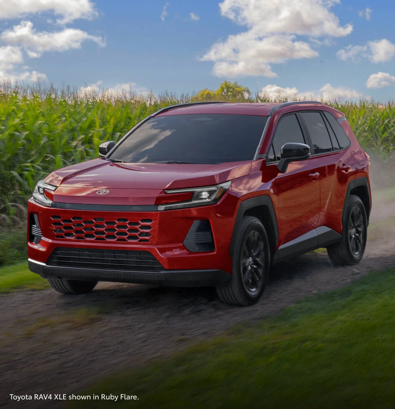 Front three-quarter view of a 2026 Toyota RAV4 XLE in Ruby Flare Pearl driving along a rural dirt road by a cornfield.