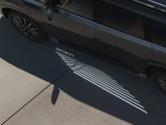 High-angle side view of a dark charcoal 2027 INFINITI QX80 LUXE projecting a bright white INFINITI welcome light carpet — wordmark and geometric wing pattern — onto the pavement.