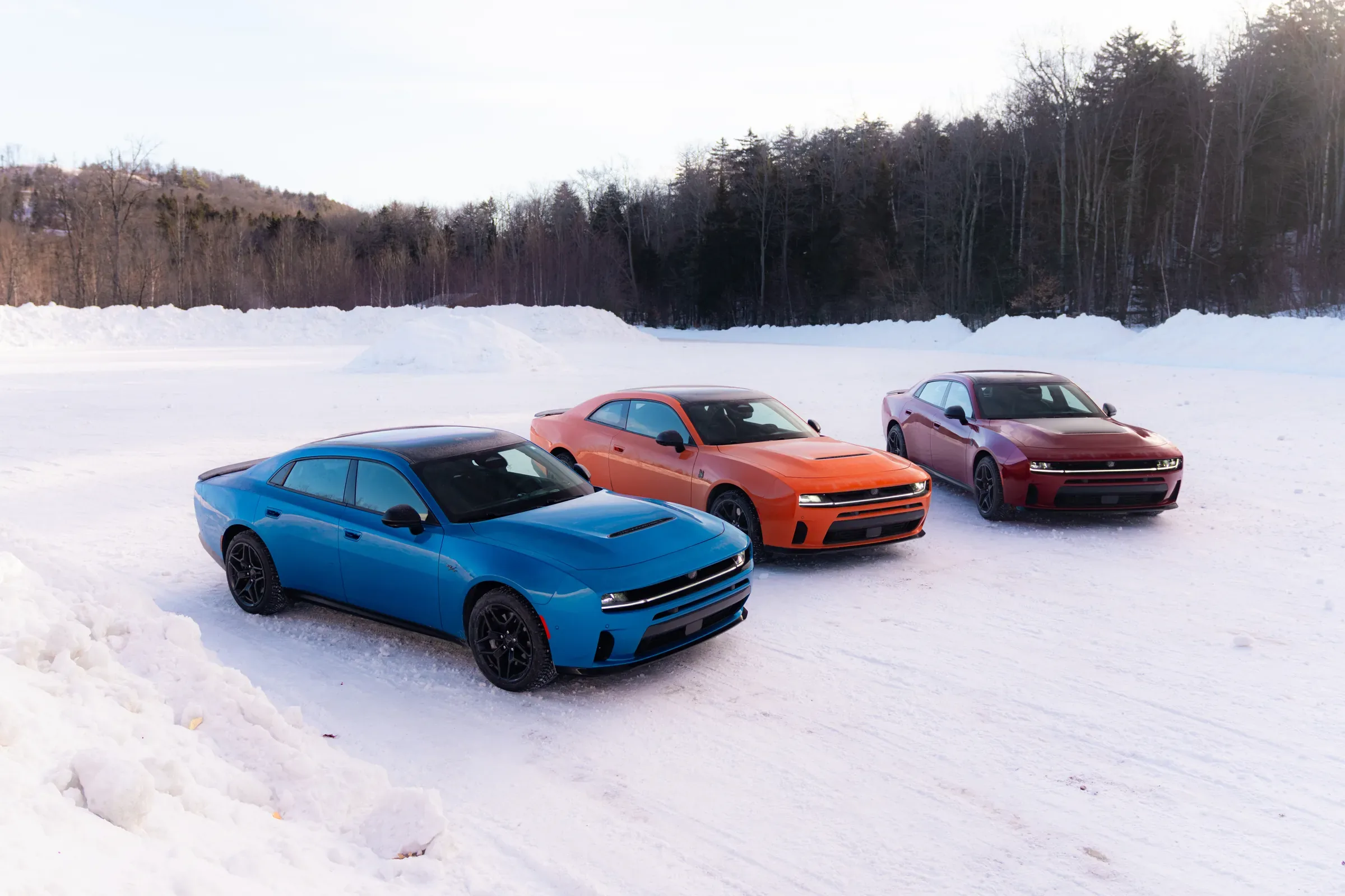 Three 2026 Dodge Chargers in blue, orange, and maroon parked together on a snow-covered field with a tree line behind them