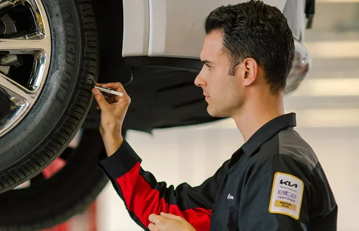 A Kia-certified technician, shown in profile, marks a tire's tread with a paint stick while inspecting it on a lift, illustrating the coverage described in the Kia Road Hazard Tire & Wheel Plan.
