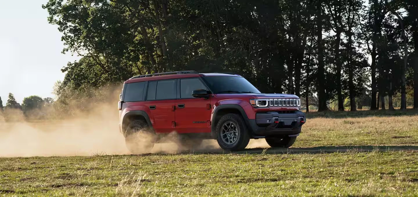 Red 2026 Jeep Recon SUV driving across a grassy field, kicking up a cloud of dust, with tall trees in the background