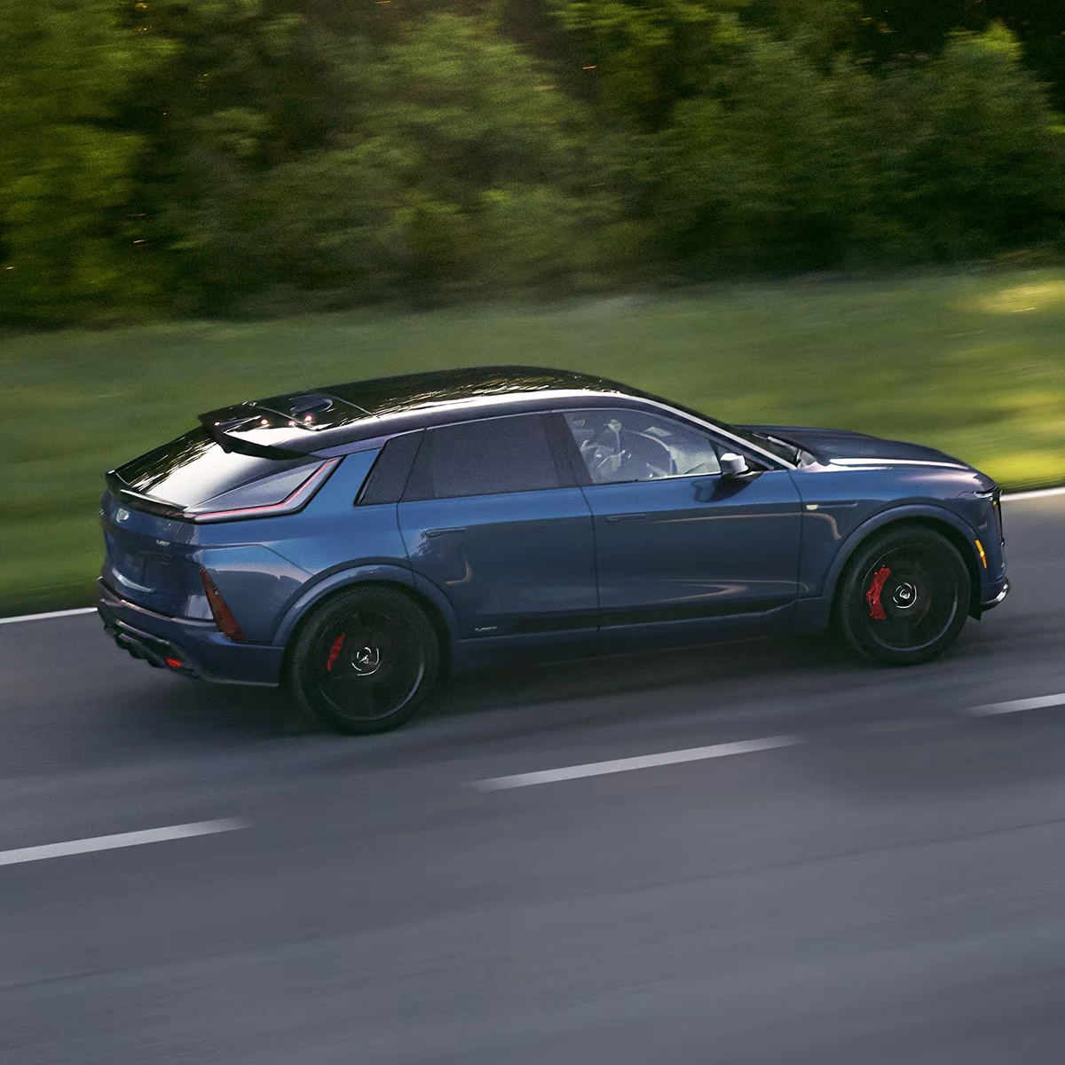 Blue 2027 Cadillac Lyriq-V SUV with a rear roof spoiler, black wheels and red brake calipers, seen from above in side profile driving along a road, with blurred green trees and grass behind it